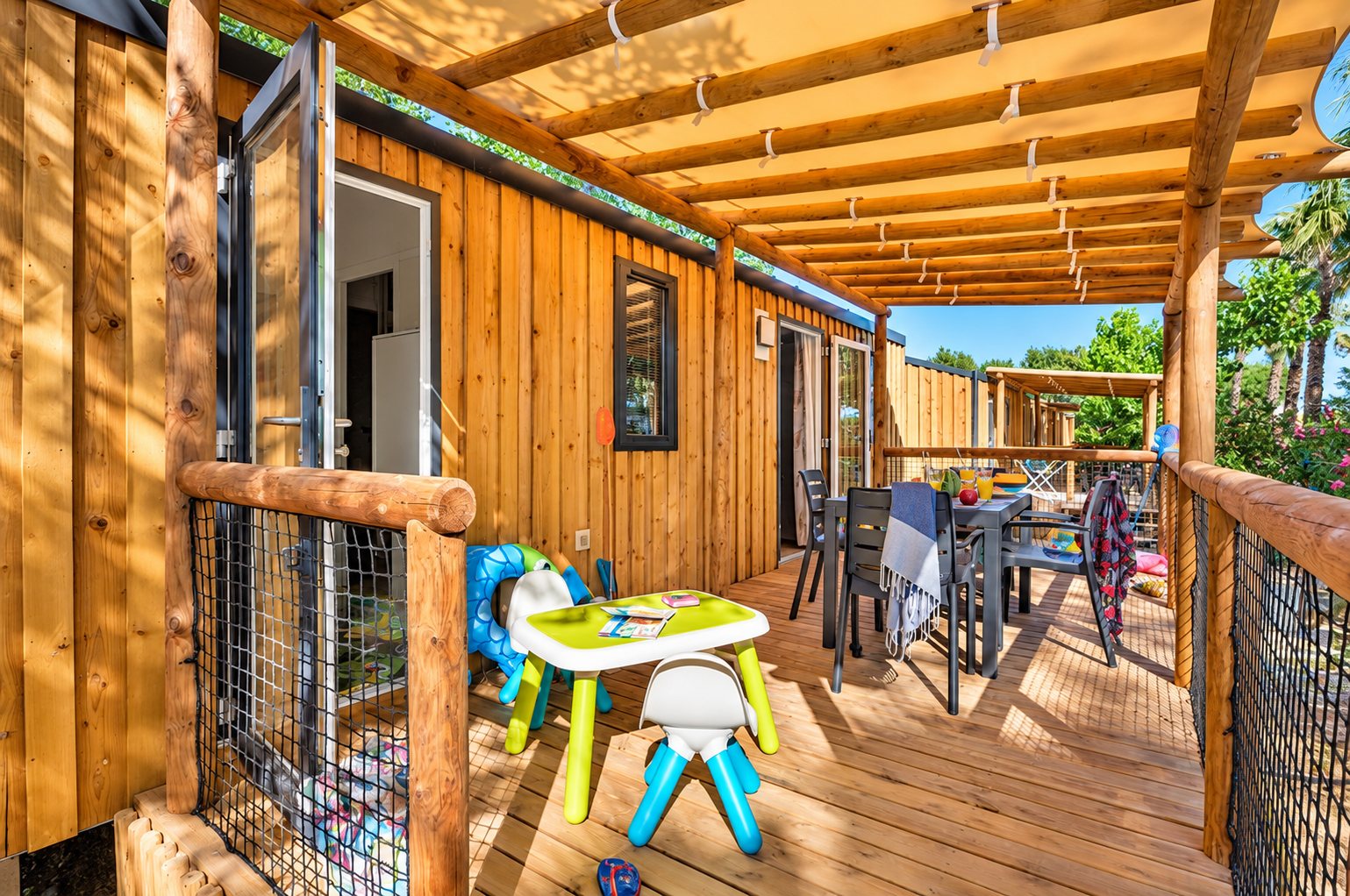 Terrasse en bois ensoleillée d’un mobil-home, avec une table et des chaises colorées pour enfants au premier plan et une table de repas au fond.