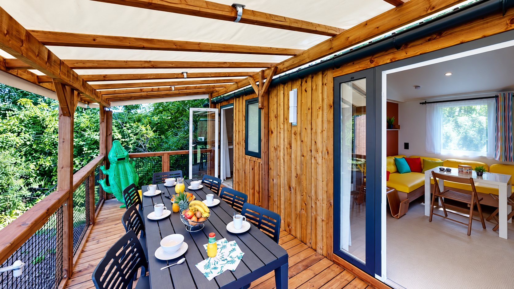 Terrasse couverte en bois d’un mobil-home avec une table dressée pour le petit-déjeuner, des fruits et des tasses, et un salon jaune visible par la porte ouverte.