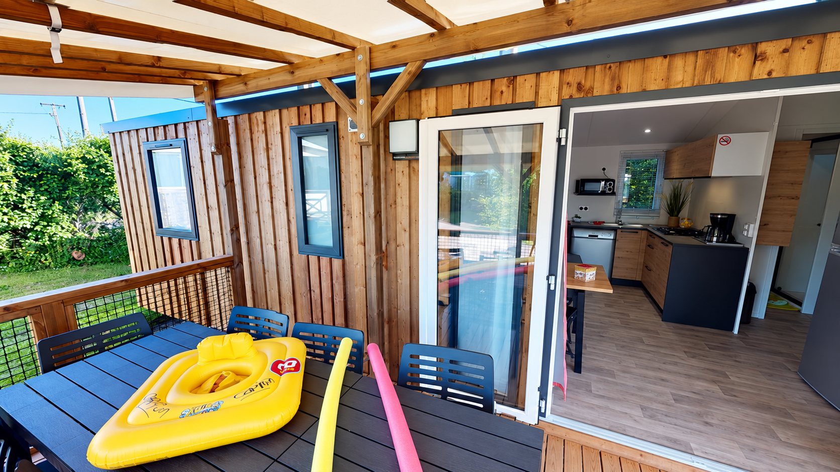 Terrasse couverte d’un mobil-home en bardage bois avec table, chaises et bouée jaune, et porte ouverte sur la cuisine équipée.